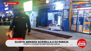 Un policía uniformado camina en una calle oscura, con una cinta de precaución amarilla y un bulto cubierto en el suelo frente a una licorería iluminada