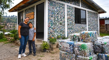 Una mujer y un niño posan al lado de una casa construida con paredes de plástico reciclado y fardos de bloques en el suelo.