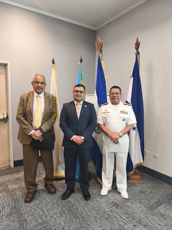 Tres hombres de pie en una oficina, dos vestidos con traje formal y uno con uniforme militar blanco, frente a banderas nacionales