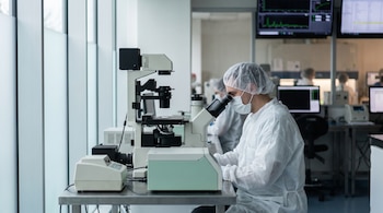 Un hombre con bata blanca, mascarilla y gorro examina una muestra en un microscopio junto a monitores de laboratorio.