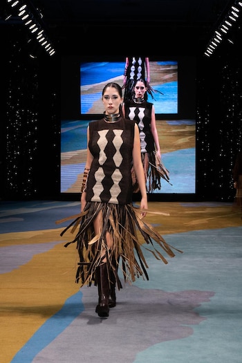 Vista frontal de dos modelos caminando por una pasarela. La modelo principal lleva un top sin mangas con patrón geométrico, una falda de tiras y botas altas