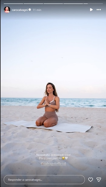 Una mujer, Caro Calvagni, está de rodillas sobre una esterilla de yoga en una playa, con las manos juntas y los ojos cerrados, frente al océano