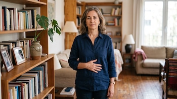 Mujer con camisa azul y la mano en el abdomen en una habitación con libreros, plantas y un sofá.