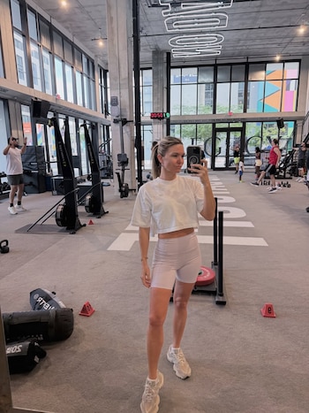 Mujer rubia de pie en un gimnasio, toma una selfi con un teléfono móvil. Se observan aparatos de ejercicio y otras personas entrenando de fondo