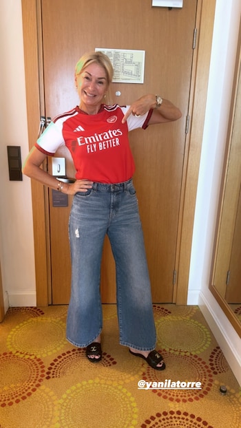 Yanina Latorre, mujer rubia de mediana edad, sonríe y señala la camiseta roja y blanca del Arsenal que viste, combinada con jeans y sandalias negras