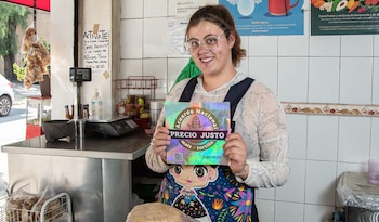 Mujer joven con gafas sonríe y sostiene un cartel plateado con el texto 'Acuerdo Nacional Precio Justo maíz - tortilla' en un mostrador de tienda