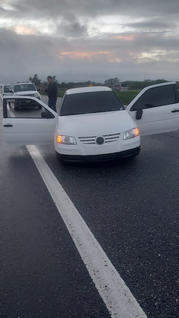 Un automóvil blanco con ambas puertas abiertas permanece sobre el asfalto mojado de una ruta, cerca de una camioneta blanca
