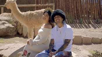 Wendy Sulca y Micro TDH sonríen sentados al aire libre, con una alpaca clara detrás. Ella lleva un vestido color crema, él un gorro peludo oscuro y camiseta blanca