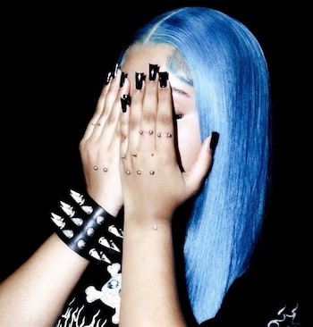 Primer plano de una persona con cabello azul brillante cubriendo su rostro con las manos. Se ven uñas negras, piercings dérmicos y una pulsera de tachuelas