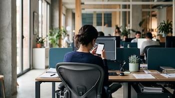 Mujer sentada en silla de oficina, vista de espalda, con teléfono móvil en mano frente a un monitor de computadora en un escritorio con plantas y papeles
