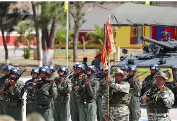 Soldados con uniformes militares, cascos y fusiles permanecen alineados junto a un estandarte rojo y un vehículo blindado