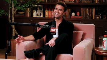 Paul Wesley sonríe sentado en un sillón rosa. Un micrófono con el logo Call Her Daddy está frente a él. Detrás hay una estantería con libros