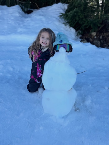 Una niña sonríe en la nieve junto a un muñeco de nieve que lleva un gorro celeste y gafas de esquí