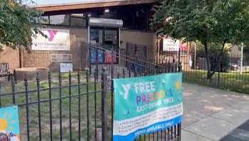 Fachada de un edificio YMCA con paredes de madera y ventanales; un cartel grande de YMCA y otro de preescolar, una valla de metal y árboles