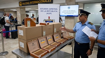 Dos oficiales de uniforme junto a cajas de puros y cartón sobre una mesa metálica en la terminal de un aeropuerto.