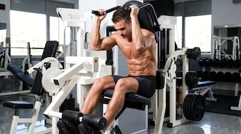Un hombre musculoso sin camiseta sentado en una máquina de gimnasio, con pesas a los lados y en el fondo, y ventanas a la izquierda.