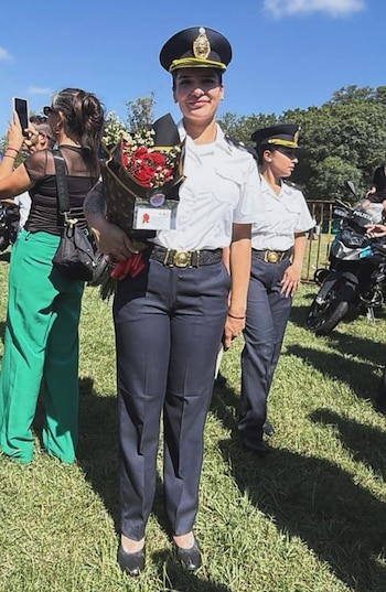Una mujer policía con uniforme azul y blanco sonríe, sosteniendo un ramo de rosas rojas, en un día soleado al aire libre