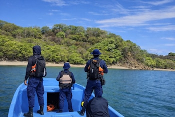 Cuatro personas con uniformes oscuros, chalecos salvavidas y mochilas en una embarcación azul observan una costa con árboles y arena.