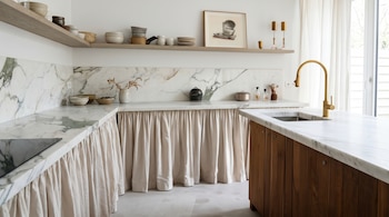 Una cocina con encimeras de mármol blanco, un faldón de tela beige bajo la mesada, estantes de madera con tazas y platos, y un grifo dorado.