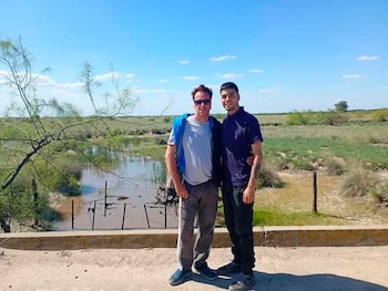 Dos hombres están de pie en un camino de tierra junto a una pequeña laguna y vegetación bajo un cielo despejado