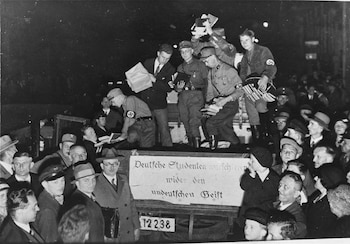 Multitud de hombres, algunos en uniformes nazis, manipulan y arrojan libros desde un vehículo en una calle oscura, con una pancarta en alemán
