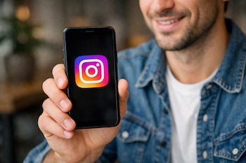 Un plano medio de una persona sonriente, vestida con una camisa de mezclilla, sosteniendo un teléfono móvil negro con el logotipo de Instagram en la pantalla.