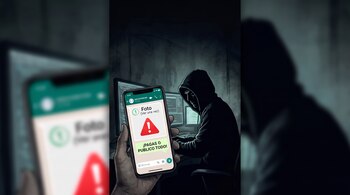 Teléfono móvil con un mensaje de texto amenazante en primer plano y una persona con capucha frente a monitores de ordenador en el fondo.