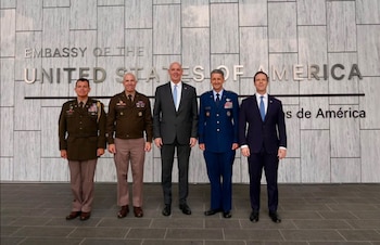 Cinco hombres de pie frente a la fachada de la Embajada de Estados Unidos con un cartel grande, dos en uniforme militar marrón, uno en uniforme militar azul y dos en traje