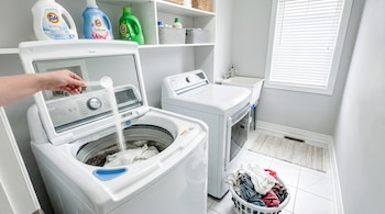 Mano que vierte detergente en una lavadora blanca abierta, junto a una secadora, un cesto con ropa y estantes con productos.