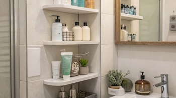 Vista de un baño moderno con azulejos claros, estantes esquineros blancos llenos de botellas y tubos de productos, un espejo de madera, un lavabo y un grifo cromado.