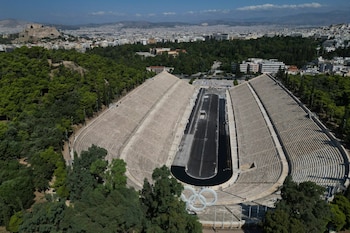 Vista aérea de un estadio con gradas de piedra, una pista oscura y un monumento de anillos blancos, rodeado de árboles, edificios y montañas