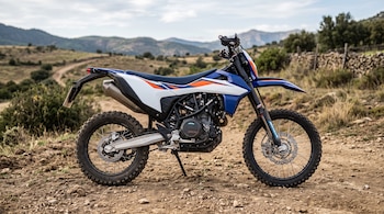 Motocicleta azul y blanca con neumáticos de tacos sobre un camino de tierra ante un paisaje de colinas.