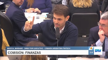 Un hombre frente a un micrófono levanta el índice durante una presentación, acompañado por un zócalo con texto explicativo