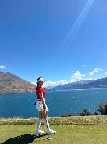 Mujer con gorra y ropa deportiva de golf en campo de césped. Sostiene un palo de golf. Al fondo hay un lago y montañas bajo un cielo azul con nubes