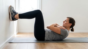 Mujer realizando un crunch abdominal en una colchoneta gris. Está acostada boca arriba, con los pies apoyados en una pared blanca y los brazos cruzados.