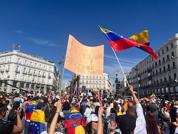 Una gran multitud de personas ondea banderas de Venezuela y pancartas en una manifestación al aire libre, bajo un cielo azul claro y edificios urbanos