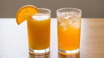 Un vaso alto con jugo de naranja y una rodaja de naranja, y otro vaso alto con gaseosa de color naranja y cubos de hielo, sobre una mesa de madera.