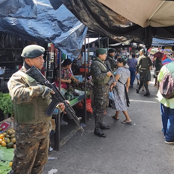 Soldados con uniforme de camuflaje y fusiles en un mercado al aire libre, con puestos de frutas, verduras y civiles comprando bajo lonas
