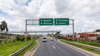 Carretera asfaltada con flechas pintadas en el suelo, un letrero verde con indicaciones hacia Bogotá y Funza, vehículos y árboles