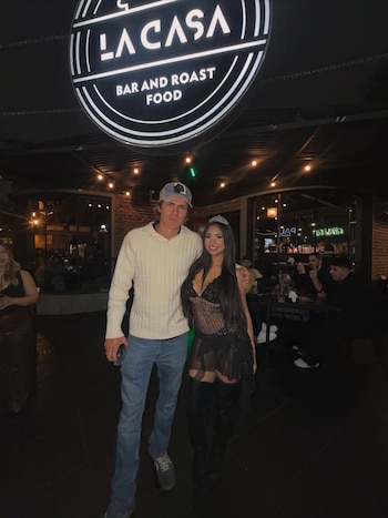Daniela Celis posa con un hombre en un restaurante nocturno. Ella lleva un vestido negro, botas altas y una tiara. Un letrero "La Casa" es visible arriba