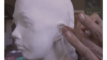 Primer plano de manos esculpiendo la oreja de una cabeza de yeso o arcilla blanca, con el perfil del rostro visible en el lado izquierdo