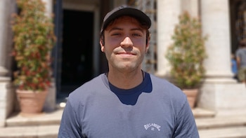 Primer plano de un joven sonriendo, con gorra oscura y camiseta gris con logo, bajo la luz del sol. El fondo borroso muestra arquitectura y plantas