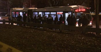 Un autobús blanco con letrero "BX5 BAY PLAZA" está rodeado por oficiales de policía de la NYPD en una calle oscura, con cinta de escena del crimen