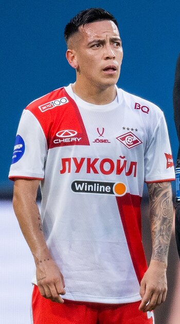 Hombre con cabello oscuro corto, con tatuajes en ambos brazos, viste una camiseta de fútbol blanca y roja con logotipos de patrocinadores