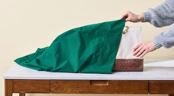 Manos de una persona cubriendo un bolso de color blanco y marrón con una funda verde sobre una mesa de mármol.