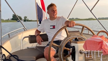 Joven rubio con camiseta blanca y pantalones cortos azules al timón de una lancha con bandera de Países Bajos y canasta con toalla naranja