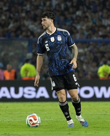 Marcos Senesi, un futbolista con cabello oscuro, de pie en un campo de césped verde, vistiendo una camiseta negra y azul con el número 6 y pantalones cortos negros, con un balón cerca