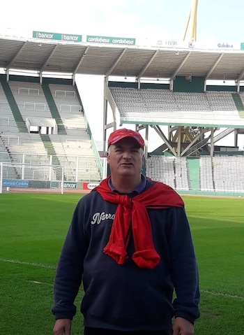 Hombre caucásico de mediana edad, Ariel Raúl Medri, con gorra roja y buzo azul, parado en el césped de un estadio de fútbol con gradas vacías