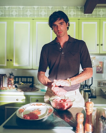 Jacob Elordi de pie en una cocina con gabinetes verdes y papel tapiz, manipulando carne picada en un tazón de vidrio. Lleva camisa oscura