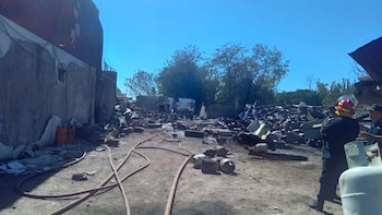 Sitio devastado por incendio en Merlo, con escombros carbonizados, estructuras parcialmente destruidas, mangueras y tanques de gas. Bomberos a la derecha bajo cielo azul